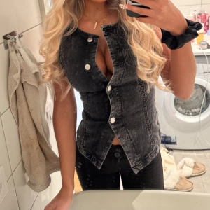 Svart jeansväst med knappar - Snygg svart jeansväst med markerade sömmar och silvriga knappar framtill. Västen har en unik asymmetrisk knäppning och är ärmlös, perfekt att styla till olika outfits. Från Gina använd 1 gång