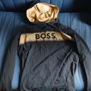 Svart och beige hoodie från BOSS - Säljer en svart hoodie från BOSS med beige panel och huva. Stor BOSS-logga framtill och kontrastfärgad insida på huvan. Perfekt för en avslappnad och stilren look.
