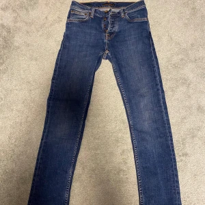 Blå jeans från Nudie Jeans - Säljer ett par klassiska blå jeans från Nudie Jeans med raka ben och orangea sömmar. Byxorna har knäppning med knappar och fem fickor. Perfekta för dig som gillar en tidlös jeansstil.