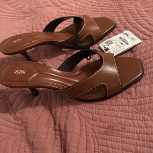 Bruna klackskor med öppen tå från Zara - Säljer ett par bruna klackskor från Zara med öppen tå och fyrkantig form framtill. Skorna har en låg klack och är tillverkade i syntetmaterial med vita kontrastsömmar. Perfekta nu inför sommaren. Klackarna har aldrig använts och prislappen är även kvar. Säljer för 300kr