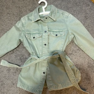 Ljusgrön jeansjacka med bälte från Zara  - Säljer en ljusgrön jeansjacka från Zara med bälte i midjan. Jackan har två bröstfickor med knappar, klassisk krage och långa ärmar. Perfekt för dig som gillar en avslappnad men trendig stil.