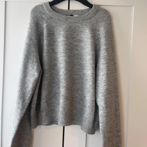 Grå stickad tröja från H&M Divided - Mysig grå stickad tröja från H&M Divided med rund halsringning och långa ärmar. Tröjan har en klassisk, enkel design och är perfekt att styla med jeans eller kjol. Ribbstickade muddar vid ärmslut och nederkant.