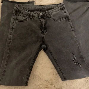 low waist med bootcut och slitning - Säljer ett par grå jeans med bootcut och en snygg slitning på ena benet. Jeansen har klassisk femficksdesign, knapp och dragkedja framtill. Perfekta för dig som gillar en lite mer avslappnad stil.