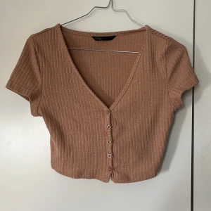 Beige ribbad croppad topp med knappar - Säljer en beige ribbad croppad topp med korta ärmar och v-ringning. Toppen har knappar framtill och är perfekt för varmare dagar. Enkel och stilren design som passar till mycket.