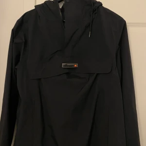 Svart vindjacka från Ellesse - Svart vindjacka från Ellesse med huva och dragkedja i halsen. Jackan har en stor ficka framtill med Ellesse-logga och meshfoder på insidan. Perfekt för blåsiga dagar och enkel att matcha med olika outfits.