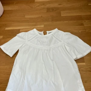 Vit blus med spetsdetaljer från Zara - Säljer en söt vit blus från Zara med fina spetsdetaljer vid axlar och halsringning. Blusen har korta ärmar och en lös passform, perfekt för en fräsch och stilren look.