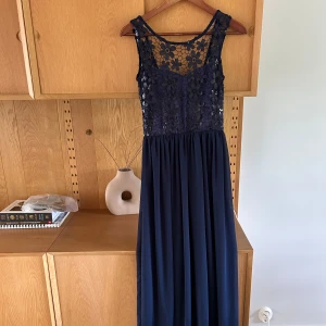 Marinblå långklänning med spets och paljetter - Säljer en marinblå långklänning med vacker spets och blommiga paljettdetaljer upptill. Klänningen har ärmlös design och en flowy kjol i mjukt tyg. Perfekt för speciella tillfällen när du vill känna dig extra fin.