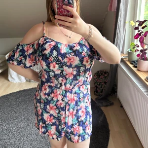 Blommig sommarplaysuit med offshoulder - Säljer en blommig playsuit med offshoulder och smala axelband. Den har ett färgglatt mönster i blått, rosa och vitt, samt volangdetaljer vid ärmarna. Perfekt för varma dagar och ger en fräsch sommarkänsla.
