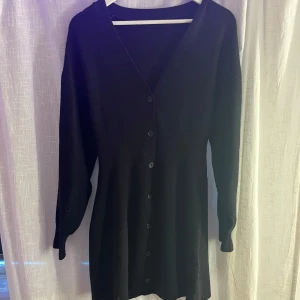 Svart stickad klänning med knappar - Fin svart klänning med lång ärm och knappar längs hela framsidan. Klänningen har en v-ringad hals och en klassisk. Aldrig gått till användning. Pris kan diskuteras.