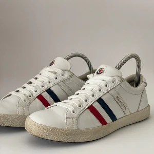 Vita sneakers från Moncler💯 - Snygga vita sneakers från Moncler med klassiska röda och blå ränder på sidorna samt Moncler-logga på plösen. Skorna har snörning och är tillverkade i skinn med platt sula och rund tå. Perfekta för en stilren och sportig look. Strl 39 men passar 40