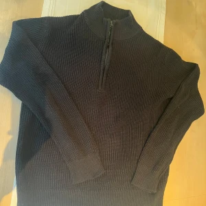 Svart stickad tröja med dragkedja från Grunt - Säljer en svart stickad tröja från Grunt i storlek XL. Tröjan har hög krage och halv dragkedja framtill, perfekt för lager-på-lager. Den är ribbstickad och har långa ärmar för extra komfort.