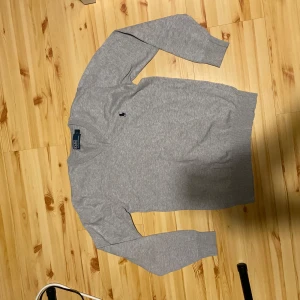 Grå tröja från Polo Ralph Lauren - Säljer en klassisk grå tröja från Polo Ralph Lauren med liten broderad logga på bröstet. Tröjan har v-ringad hals och långa ärmar. Perfekt för en stilren och enkel look.
