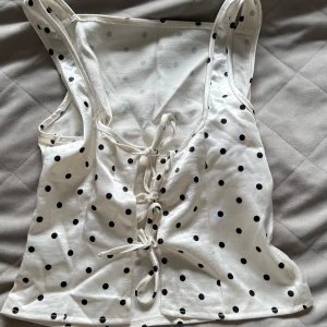 Vit prickig linnetopp med knytning bak  - Säljer en söt vit linnetopp med svarta prickar och knytning baktill. Toppen har smala axelband och en figurnära passform. Perfekt för varma dagar och enkel att matcha med jeans eller kjol.