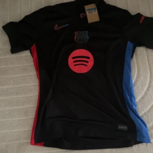 FC Barcelona fotbollströja Lamine Yamal 19 - Säljer en svart FC Barcelona fotbollströja med röd och blå detalj på sidorna. Tryck med Spotify-logga fram och Lamine Yamal 19 bak. Kortärmad modell med klubbmärke och Nike-logga på bröstet.