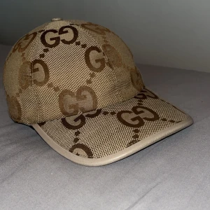 Beige keps från Gucci med GG-mönster - Säljer en beige keps från Gucci med det klassiska GG-mönstret i brunt. Kepsen har böjd skärm och är tillverkad i ett canvas tyg. Äkta