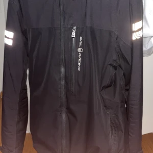 Svart vindjacka från Sail Racing - Svart vindjacka från Sail Racing med dragkedja framtill och reflekterande detaljer på ärmarna. Jackan har en bröstficka med dragkedja och är tillverkad i ett vattenavvisande material. Perfekt för blåsiga dagar jackan är storlek XL och den är bra skick förutom att de finns ett litet hål där back på jackan annars e den i jätte bra skick