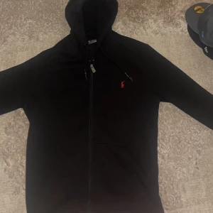 Svart hoodie från Polo Ralph Lauren - Svart hoodie med dragkedja från Polo Ralph Lauren. Klassisk design med röd broderad logga på bröstet och justerbar huva. Perfekt för en avslappnad och stilren look.