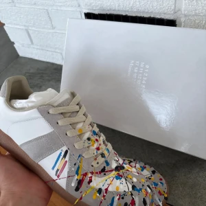 Maison Margiela sneakers med färgstänk - Unika sneakers från Maison Margiela med vit bas, grå mockadetaljer och färgstänk i blått, rött, gult och svart. Skorna har snörning, rund tå och en brun gummisula. Perfekt för dig som vill sticka ut med din stil.