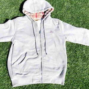 Grå zip-hoodie från Burberry - Säljer en grå hoodie med dragkedja från Burberry. Jackan har klassiskt rutigt foder i huvan och broderad logga på bröstet. Mjuk bomullskänsla och två fickor framtill. Perfekt för en chill och stilren look.