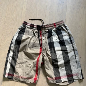 Badshorts med burberry mönster  - Shorts med stort rutigt mönster i beige, svart, vitt och röda detaljer. De har elastisk midja med snörning och är tillverkade i ett lätt syntetmaterial. Perfekta för sommaren och har en avslappnad passform. Storleken är 5XL men de sitter som L/XL