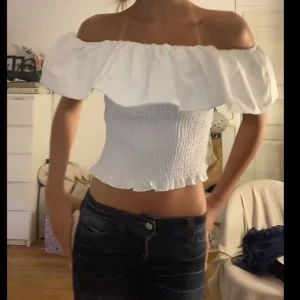 Vit off shoulder tröja - Säljer en vit offshoulder topp med volangkant. Toppen är croppad och har en smockad, tight passform som sitter snyggt på kroppen. Perfekt för sommardagar eller när du vill ha en luftig och trendig look.