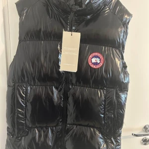 Svart dunväst från Canada Goose - Säljer en glansig svart dunväst från Canada Goose med klassisk logotyp på bröstet och dragkedja framtill. Västen har stora fickor och hög krage, perfekt för kyliga dagar. Märkeslapp på ryggen och autenticitetsmärkning på insidan.