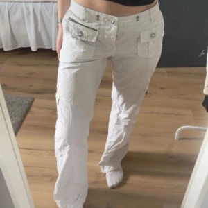 Vita cargopants med fickor och knappar - Säljer ett par vita cargopants med stora fickor framtill och bak, samt dekorativa knappar. Byxorna har en normal passform och raka ben. Nedersta knappen saknas men ingenting som syns eller märks , annars jättefint skick!