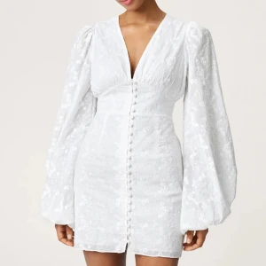 Vit klänning från Adoore - Supersnygg klänning från Adoore med v-ringning, långa puffärmar och knappar framtill. Klänningen har ett subtilt blommigt mönster och markerad midja. Perfekt för sommarens alla tillfällen!