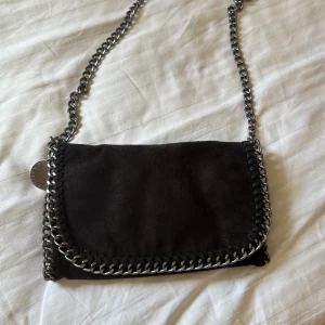 Stella McCartney liknande väska - Säljer en svart axelväska med kedjerem och dekorativa kedjedetaljer runt kanterna. Väskan har lockstängning med magnet och är tillverkad i syntetmaterial med mockakänsla. Perfekt för att lyfta din outfit! Den är inte äkta men är väldigt lik Stella McCartneys väska. Hör gärna av er vid intresse eller om ni har några frågor!🥰