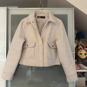 Beige croppad jacka från Zara - Säljer en beige croppad jacka från Zara med klassisk krage och stora fickor framtill. Jackan har långa ärmar, knappar och är tillverkad i ett ull-liknande material. Perfekt för lager-på-lager-stil under kyligare dagar men även perfekt nu till sommaren. Använd en gång så den är som ny och har inga defekter. Pris kan diskuteras 