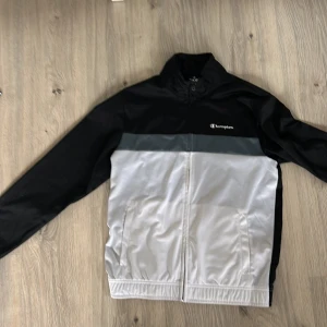 Svart och vit vindjacka från Champion - Säljer en snygg vindjacka från Champion i svart och vitt med grå detalj över bröstet. Jackan har dragkedja framtill, ståkrage och två sidofickor. Perfekt för vår och höst när det blåser lite extra.