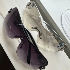 Solglasögon - Två par vintage solglasögon med futuristisk design. Ena paret har mörkbruna, nästan svarta glas och det andra har ljusbeige, transparenta glas. Båda har metalliska detaljer vid sidorna och saknar tydliga bågar runt glasen. 1 för 60kr. 2 för 100kr