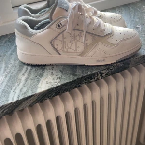 Vita Dior sneakers med monogram - Säljer ett par vita Dior sneakers med grå detaljer och klassiskt Dior-monogram på sidan. Skorna har snörning, perforerad tå och platt sula. Perfekta för dig som gillar exklusiva och stilrena sneakers.