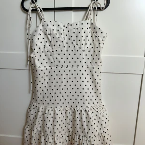 Vit prickig klänning med volang - Söt vit klänning med svarta prickar och volang nertill. Axelbandsmodell med knytbara axelband och figurnära passform upptill. Perfekt för sommarens alla tillfällen! 