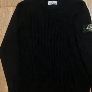 Svart stickad tröja från Stone Island - Säljer en svart stickad tröja från Stone Island med rund halsringning och klassisk logotyp-patch på vänster ärm. Tröjan har lång ärm och ribbade muddar för en snygg och enkel look.