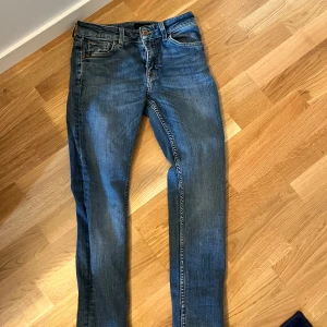 Blå jeans från Tiger of Sweden - Säljer ett par klassiska blå jeans från Tiger of Sweden med raka ben och snygga detaljer. Jeansen har fem fickor, bruna sömmar och en läderpatch med logga bak i midjan. Tillverkade i italienskt premiumdenim.