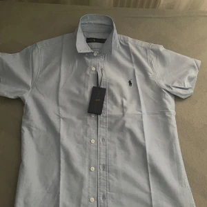 Ljusblå kortärmad skjorta från Ralph Lauren - Snygg ljusblå skjorta från Ralph Lauren med klassisk krage och korta ärmar. Skjortan har knappar framtill och den ikoniska broderade loggan på bröstet. Perfekt för en stilren look.