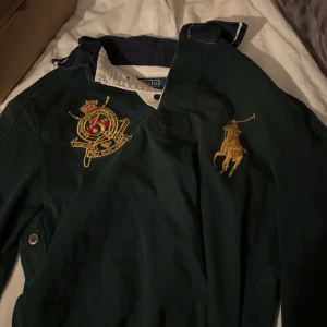Mörkgrön jacka från Polo Ralph Lauren - Säljer en mörkgrön tröja från Polo Ralph Lauren med broderade detaljer på bröstet, inklusive den klassiska polospelaren och ett emblem. Tröjan har krage i kontrastfärg och knäppning framtill. Perfekt för dig som gillar sportig och preppy stil. Den är äkta och är i storlek S.