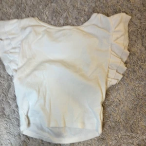 Vit topp med volangärmar - Säljer en söt vit topp med volangdetaljer på ärmarna. Toppen har en rund halsringning och är tillverkad i ett mjukt material. Perfekt för dig som gillar en feminin och stilren look.
