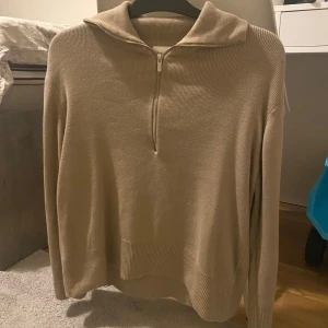 Beige stickad tröja med halv dragkedja - Säljer en beige stickad tröja med krage och halv dragkedja framtill. Tröjan har lång ärm och ribbade muddar vid ärmslut och nederkant. Perfekt för lager-på-lager och enkel att matcha med olika outfits. Använt ett par gånger. Pris kan diskuteras.