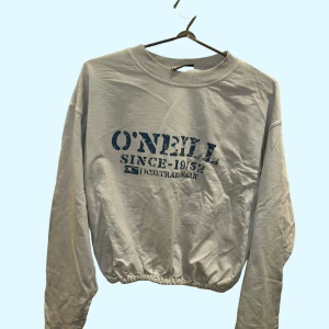 Grå croppad sweatshirt från O'Neill - Säljer en ljusgrå croppad sweatshirt från O'Neill med blå tryck framtill. Tröjan har långa ärmar och resår i nederkanten för en snygg passform. Perfekt till jeans eller mjukisbyxor.