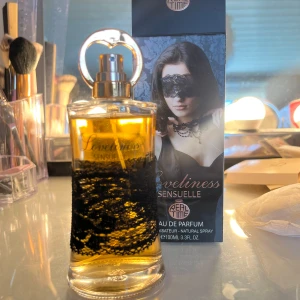Loveliness Sensuelle Eau de Parfum - En elegant parfymflaska med svart spetsdetalj och guldtonad vätska. Förpackningen har en mystisk design med en kvinna i svart spetsmask och smycken. Innehåller 100 ml Eau de Parfum och utstrålar en sensuell och lyxig känsla.
