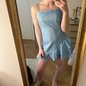 Blå blommig kortklänning med volang och öppen rygg - Supersöt blå kortklänning med vita småblommor och volangkant nertill. Klänningen har smala axelband, omlottkänsla och en snygg knytning i ryggen som ger en öppen rygg. Perfekt för sommaren! Nypris 700kr.