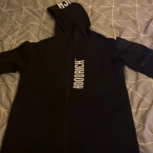 Svart hoodiejacka från Hoodrich - Svart hoodiejacka från Hoodrich med vit logga tryckt längs dragkedjan och på huvan. Jackan har lång ärm och dragkedja framtill. Perfekt för en streetwear-look.