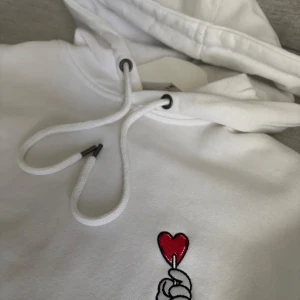 Vit hoodie med broderat hjärta - Säljer en vit hoodie med broderad detalj på bröstet – en hand som håller ett rött hjärta. Tröjan har huva med dragsko och en stor magficka. Perfekt för dig som gillar enkel men snygg stil. Köpt för 599kr