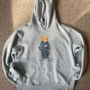Grå polo bear hoodie - Säljer en grå hoodie från Ralph Lauren med det klassiska Polo Bear-motivet framtill. Tröjan har huva med dragsko och är tillverkad i mjuk bomullsblandning. Perfekt för en avslappnad och stilren look. Extremt bra skick, köpt för inte så längesen och använt sparsamt, priset är som alltid diskuterbart mina vänner, skriv om ni har frågor❤️🥂