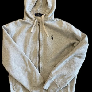 Grå hoodie från Polo Ralph Lauren - Snygg ljusgrå hoodie från Polo Ralph Lauren med dragkedja och huva. Klassisk broderad logga på bröstet och praktiska fickor framtill. Perfekt för en avslappnad stil.