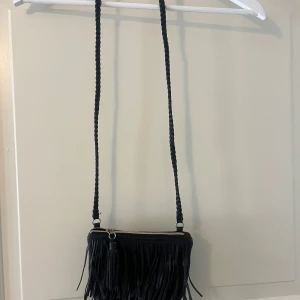 Svart axelväska med fransar - Säljer en svart axelväska med långa fransar och flätat axelband. Väskan har dragkedja upptill och ett vinrött innerfoder. Använder inte.💕