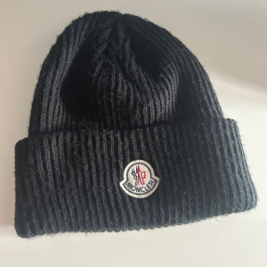 Svart ribbad mössa från Moncler - Säljer en svart ribbstickad mössa från Moncler med uppvikt kant och broderad Moncler-logga framtill. Klassisk och stilren design som passar perfekt till kyliga dagar.