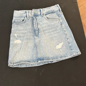 Ljusblå jeanskjol från h&m - Säljer en ljusblå jeanskjol från h&m med klassisk femficksdesign och slitna detaljer framtill. Kjolen har bälteshällor och är i en kort modell som passar perfekt till vardags. Enkel att matcha med olika toppar.